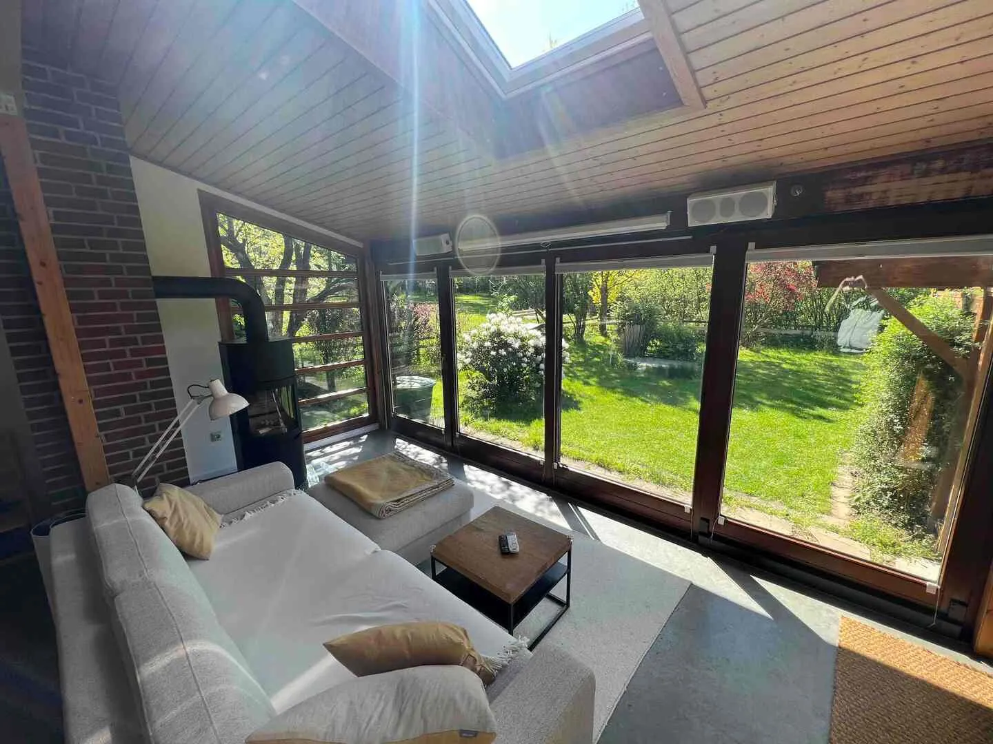 Sonne fällt durchs Dachfenster in den Wohnraum: helles Sofa vor der Fensterfront, dahinter der grüne Garten, links Backsteinwand und Kaminofen
